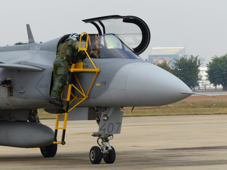 A pilot in a flight suit climbs into the cockpit of a military jet on the runway.