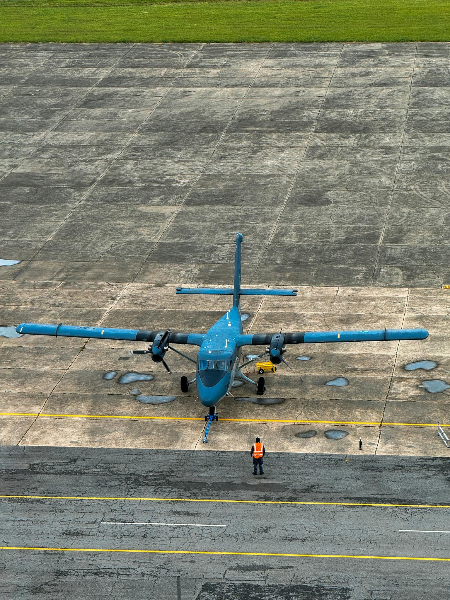 A small blue twin-propeller airplane parked outdoors with a ground crew member visible.